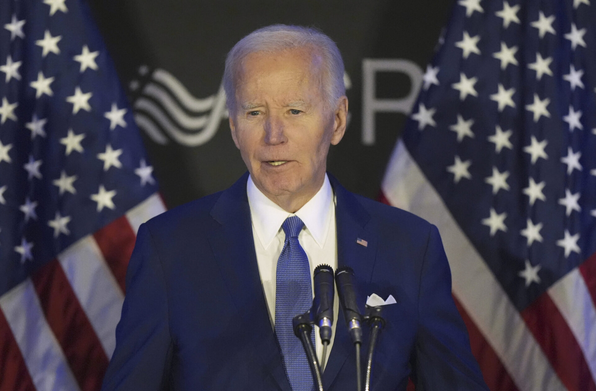 Former President Joe Biden speaks at a podium in a suit and tie, looking resolute during an outdoor event.