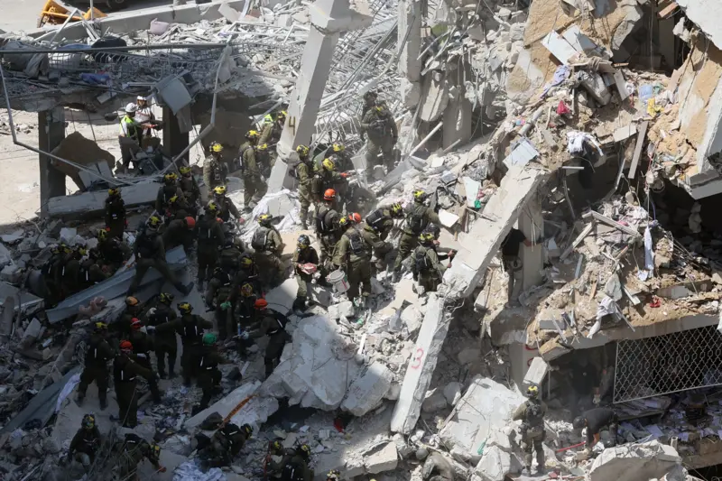 The entire wing of the residential complex has collapsed, with twisted metal rods and cracked concrete slabs hanging precariously from the remains.